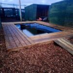 A long view between shed like structures. In the background are high mesh fences predator proofing the enclosure. In the centre of the image is large pool with wooden decking around the edges. There is woodchip on the floor.